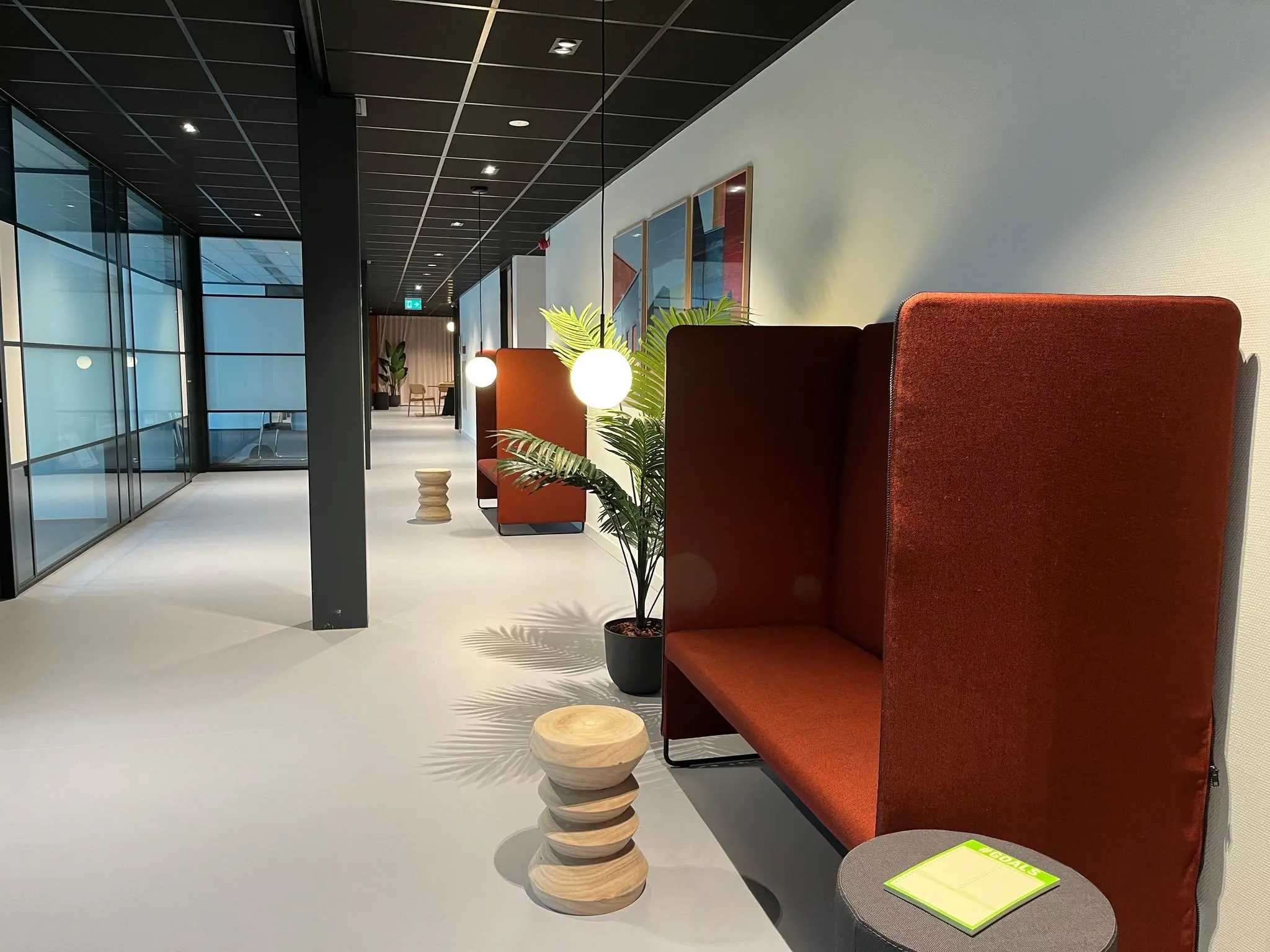 Moderne kantoorruimte met roodbruine zitnissen, houten krukjes, planten en sfeervolle verlichting langs een glazen gang.